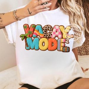 Op de afbeelding: Wit T-shirt met de tekst "VACAY MODE" in kleurrijke, getextureerde letters. Het ontwerp bevat palmbomen, een luipaardprint, een roze en wit geblokt patroon en een kleine koffer, wat een vakantiethema oproept.