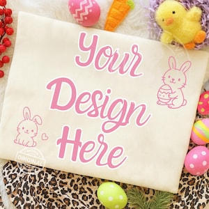 以下が含まれることがあります： ピンク色の「Your Design Here」の文字が入ったベージュ色のトートバッグ。バッグにはピンク色のウサギと卵のイラストが描かれています。卵やひよこなど、イースターをテーマにした装飾がバッグを囲んでいます。