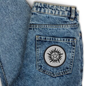 Könnte beinhalten: Blaue Acid-Wash-Jeans mit einem runden gestickten Aufnäher auf der Gesäßtasche. Der Aufnäher hat einen silbergrauen Hintergrund mit schwarzem Rand und ein zentrales schwarzes Pentagramm-Design, das von stilisierten flammenartigen Sonnenstrahlen umgeben ist.