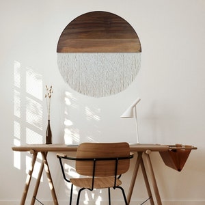 May include: A wooden and white macrame wall hanging in the shape of a half circle. The wall hanging is hanging on a white wall above a wooden desk with a chair.