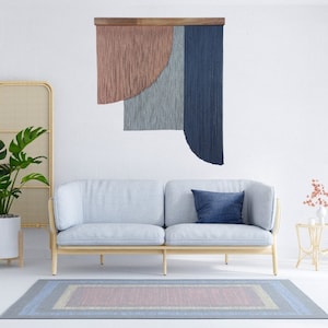 May include: A light blue sofa with a blue pillow sits in front of a wall tapestry with pink, blue, and gray yarn fringe. A small table with a vase of pink flowers sits to the right of the sofa. A rug with a pink, blue, and gray pattern is on the floor.