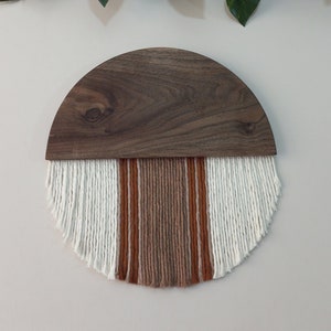 May include: A half-circle wooden wall hanging with a fringe of white, brown, and orange yarn.