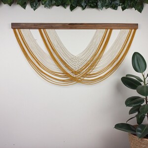 May include: A macrame wall hanging with a white and yellow colour scheme. The hanging is made of yarn and is attached to a wooden dowel.