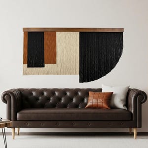 May include: A contemporary wall hanging with a wooden frame. The artwork displays geometric shapes in black, tan, and cream. A brown leather sofa with buttoned cushions is positioned below. Decorative cushions in white and brown complete the scene.