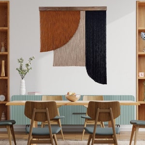 Op de afbeelding: Een abstracte wandhanger met een houten frame. Het kunstwerk toont secties van roest, beige en zwarte getextureerde vezels. Het stuk hangt boven een houten eettafel met stoelen en creëert een modern, artistiek interieurontwerp.
