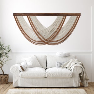 May include: A white macrame wall hanging with brown leather accents. The hanging is in the shape of a crescent moon and is mounted on a wooden dowel.