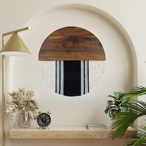 May include: A round wall hanging with a wooden top half and a white and black striped bottom half. The wooden top is a dark brown color and the stripes are made of yarn.