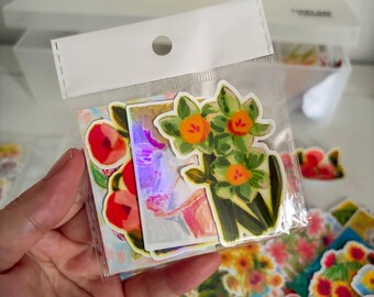 May include: A clear plastic bag containing several die-cut stickers featuring colourful floral illustrations. The visible stickers include a cluster of yellow and green daffodils, a red flower, and a rectangular sticker with a purple and pink abstract design.