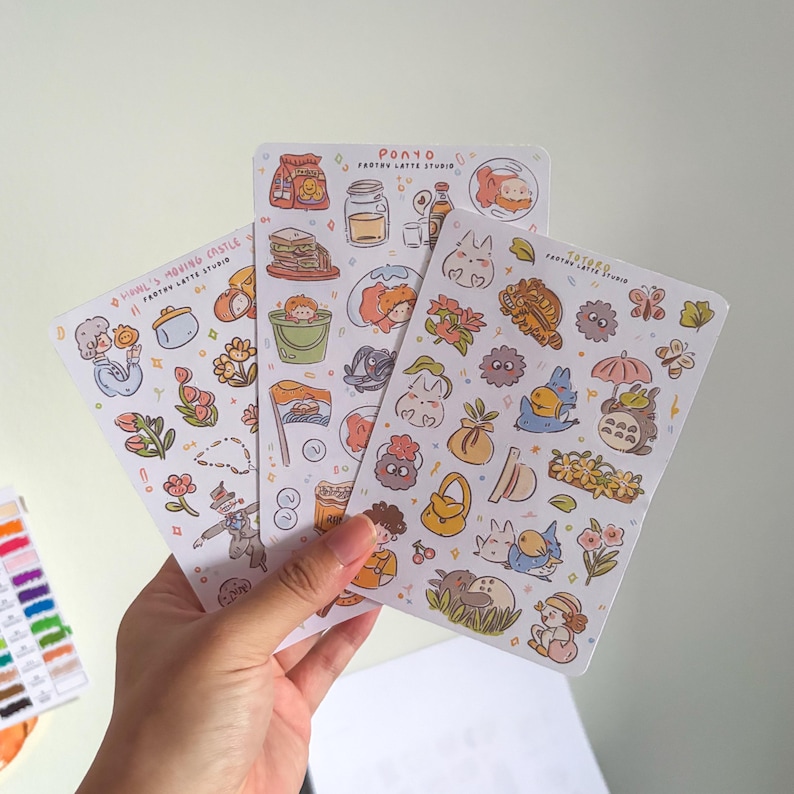 Puede incluir: Tres hojas de pegatinas con ilustraciones coloridas de personajes y objetos animados. Las hojas incluyen im&aacute;genes de comida, flores y varias figuras de dibujos animados. El texto "Ponyo Frothy Latte Studio" y "Howl's Moving Castle" son visibles.