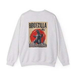 Puede incluir: Sudadera gris claro con un gr&aacute;fico en la espalda. El gr&aacute;fico presenta un monstruo frente a un sol rojo, con la palabra "BRIDEZILLA" arriba. Tambi&eacute;n se ve un helic&oacute;ptero y el horizonte de una ciudad.