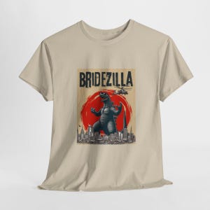 May include: A beige t-shirt with a graphic print of a Godzilla-like monster in front of a cityscape. The text "Bridezilla" is printed above the monster.