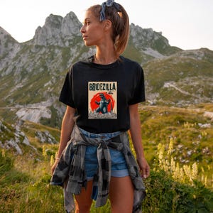 May include: A black t-shirt with a vintage-style graphic of a Godzilla-like monster with the text "Bridezilla" above it. The person wearing the shirt is standing in a mountainous landscape.