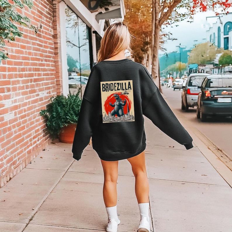 Puede incluir: Sudadera negra con un gr&aacute;fico en la espalda. El gr&aacute;fico presenta el texto "BRIDEZILLA" sobre una ilustraci&oacute;n de un monstruo frente a un sol rojo y un horizonte urbano. La sudadera la lleva una persona en una acera.