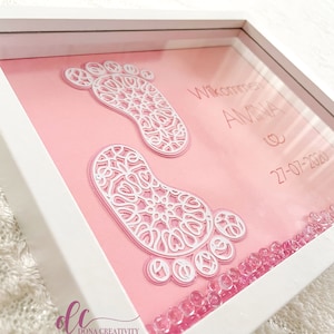 May include: A framed baby keepsake featuring two white, intricately designed footprints on a pink background. The frame includes the name "AMINA" and the date "27-07-2020". Pink beads are at the bottom of the frame.