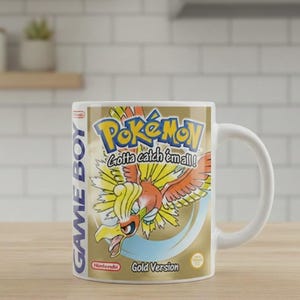 Puede incluir: Una taza de cerámica blanca con la ilustración de la portada del videojuego de Nintendo Game Boy Pokemon Edición Oro. El diseño incluye al Pokemon Ho-Oh, el texto Pokemon Gotta catch 'em all!, el logo de Game Boy y el logo de Nintendo en dorado.
