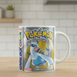 Puede incluir: Una taza de cerámica blanca con la ilustración de la portada del juego Pokémon Edición Plata para Game Boy Color. El diseño incluye al Pokémon Lugia, el logotipo de Pokémon, el texto Gotta catch 'em all!, el logo de Nintendo y la marca Game Boy Color.