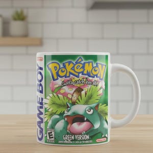 以下が含まれることがあります： 任天堂ゲームボーイ用ソフト「ポケットモンスター 緑」のパッケージアートがプリントされた白いセラミック製マグカップ。緑色の背景に、ポケモンロゴ、Gotta catch 'em all!の文字、フシギバナのキャラクター、ESRB Eレーティングシンボルがデザインされています。