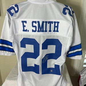 May include: White football jersey with blue trim and the number 22 on the back. The name "E. SMITH" is printed above the number. The jersey is signed in blue ink.