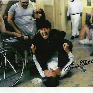 May include: A signed photograph featuring two men in a dramatic pose. One man is on top, wearing a black jacket, while the other is on the ground in a white shirt. Other people are in the background. The signature "Louis Fletcher" is visible.