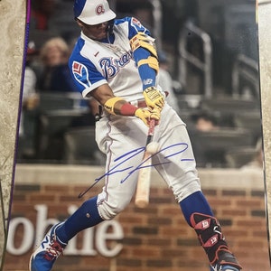 May include: A signed photograph of a baseball player in action, swinging a bat. The player is wearing a white, blue, and red uniform with the word "Braves" across the chest. The photo is signed in blue ink.
