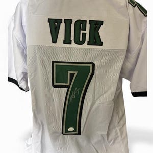 Könnte beinhalten: Weißes Football-Trikot aus Mesh mit dem Namen "VICK" in grünen Blockbuchstaben auf dem Rücken. Die Nummer "7" ist ebenfalls grün mit schwarzer Umrandung, und eine Unterschrift ist sichtbar. Das Trikot hat schwarze Verzierungen.