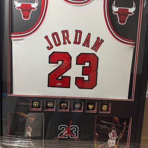 以下が含まれることがあります： 赤い文字で「JORDAN 23」と書かれた、額装されたシカゴ・ブルズのジャージ。ジャージは白地に赤と黒のトリムで、サイン入り。フレームには、ブルズのロゴ、チャンピオンシップリング、アクション写真も含まれています。