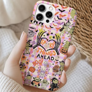 May include: A phone case with a Halloween theme. The case has a pink and purple checkered pattern with spooky illustrations, including bats, skeletons, and pumpkins. The text "Spooky Things Ahead" is displayed on the case.