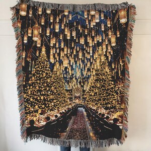 May include: A woven throw blanket depicting a grand hall with floating candles and a long dining table. The scene is lit with warm, golden light against a dark blue backdrop. The blanket has a fringed edge and is suitable for a bed or sofa.