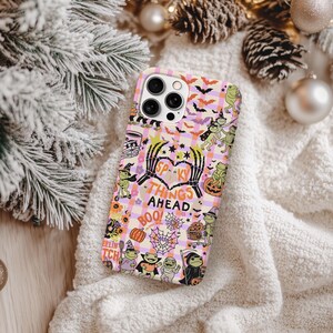 May include: A phone case with a Halloween theme. The case features a pink and orange checkered pattern with bats, frogs, pumpkins, and the text "Spooky Things Ahead." The design includes a heart made of skeleton hands.