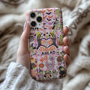 May include: A Halloween phone case with a plaid design, decorated with bats, pumpkins, and costumed frogs. The case features the text "Spooky Things Ahead" and "Boo!" in a fun font.