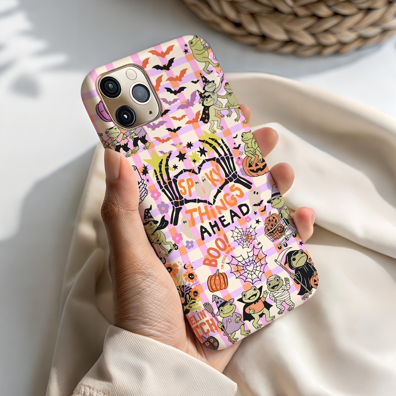 May include: A phone case with a Halloween theme. The case features a pink and white checkered pattern with bats, frogs, pumpkins, and the text "Spooky Things Ahead!" in a heart shape. The case has a glossy finish.