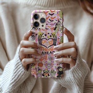 May include: A phone case with a Halloween design. The case has a pink and yellow plaid background with bats, pumpkins, spiders, and cartoon characters. The text "Spooky Things Ahead" and "Boo!" are also visible.