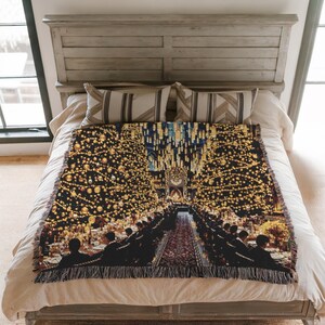 May include: A woven throw blanket depicting a grand hall scene with long tables and floating candles. The blanket has a black backdrop with gold and yellow detailing, and a fringed border. The blanket is displayed on a bed, with a neutral colour scheme.