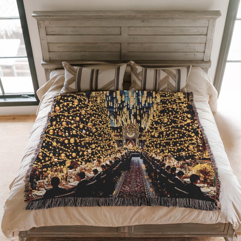 May include: A woven throw blanket depicting a grand hall scene with long tables and floating candles. The blanket has a black backdrop with gold and yellow detailing, and a fringed border. The blanket is displayed on a bed, with a neutral colour scheme.