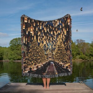 May include: A woven throw blanket depicting a grand hall with long tables, illuminated by hanging lanterns and Christmas trees. The blanket has a black background and a fringed edge. The scene is reminiscent of a magical setting.