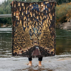 May include: A woven throw blanket depicting a festive hall with Christmas trees and hanging lanterns. The blanket has a dark background with gold and yellow accents, and a fringed edge.