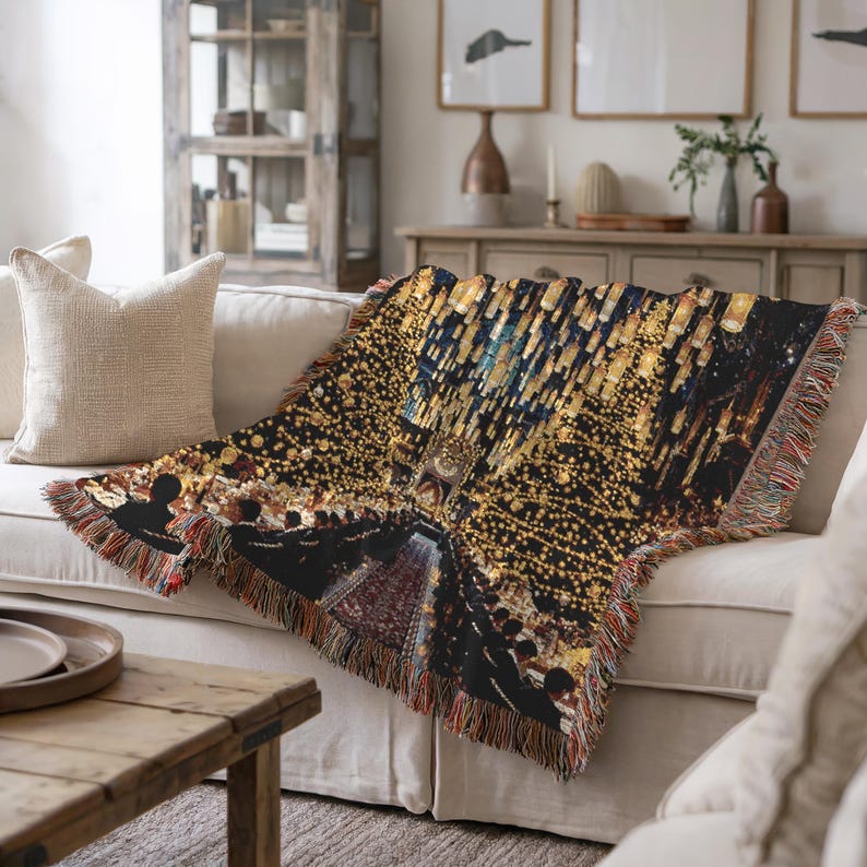 May include: A woven throw blanket featuring a city lights design in gold, blue, and black, draped over a cream sofa. The blanket has a fringed edge with red, brown, and tan threads. A decorative home accent.