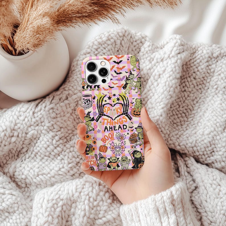May include: A Halloween-themed phone case with a pink and white checked pattern. The case is decorated with bats, pumpkins, and cartoon characters. The text "Spooky Things Ahead" is visible, adding to the festive design.