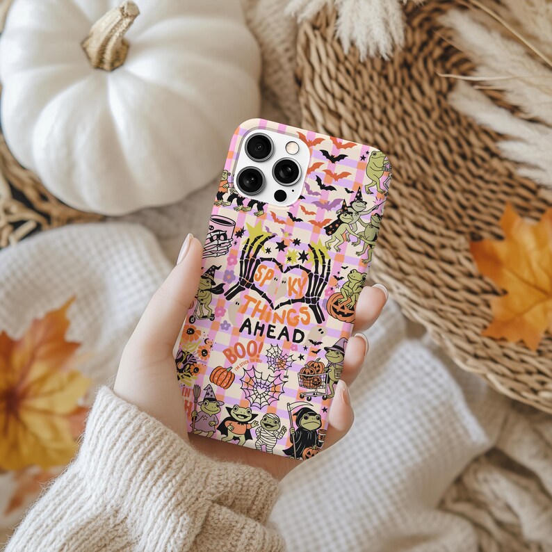 May include: A phone case with a Halloween theme. The case has a pink and purple checkered pattern with spooky illustrations, including bats, skeletons, and pumpkins. The text "Spooky Things Ahead" is displayed on the case.