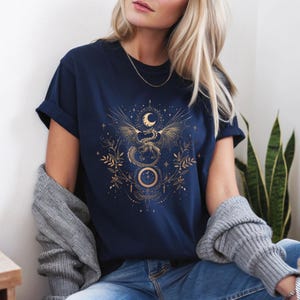 May include: A navy blue t-shirt featuring a gold graphic of a dragon with outstretched wings, a crescent moon, and stars. The design also incorporates floral elements and geometric shapes.