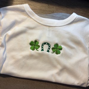 May include: White crew neck t-shirt with green embroidered St. Patrick's Day design. The design includes two four-leaf clovers and a horseshoe. The shirt is folded and ready to wear.