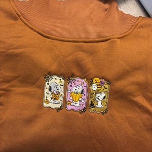 May include: A burnt orange sweatshirt featuring a ribbed mock neck collar and three embroidered rectangular panels. Each panel depicts the character Snoopy in Halloween-themed scenes with pumpkins, ghosts, spiderwebs, and bats in shades of yellow, purple, and white.