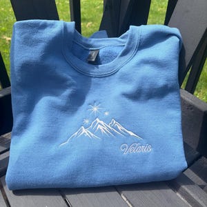 May include: A light blue crewneck sweatshirt featuring white embroidery of a mountain range topped with three starbursts. The word Velaris is embroidered in a white cursive script font below the mountain design on the front of the garment.