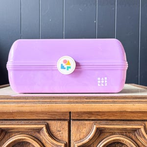 May include: A lavender-colored, rectangular lunchbox with a rounded top and a white circular emblem. The emblem features colorful geometric shapes. The text "OAB LLE" is printed on the side of the lunchbox.