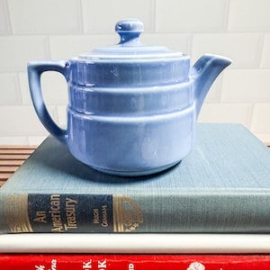 May include: A light blue ceramic teapot with a glossy finish, featuring a rounded body with three horizontal ridges, a matching lid with a small knob, a curved handle, and a short spout, resting on a stack of three hardcover books.