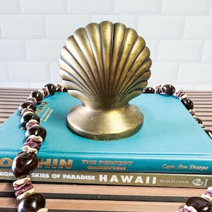 May include: A brass-toned metal shell sculpture with a ribbed surface sits atop two stacked books. The top book is teal with the text "DOLPHIN THE PERFECT GAMEFISH Capt Jim Sharpe" and the bottom book is titled "SKIES OF PARADISE HAWAII".