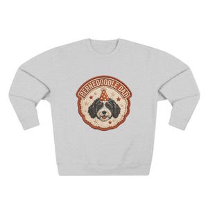 May include: A light gray long-sleeve sweatshirt featuring a circular graphic on the chest. The graphic shows a black and white Bernedoodle dog wearing a polka-dot party hat, surrounded by stars and the text "BERNEDOODLE DAD" in orange letters.