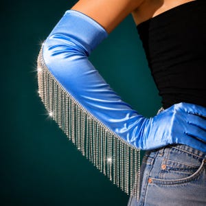 May include: A bright blue satin opera-length glove featuring a long fringe of sparkling clear rhinestones attached along the outer arm. The smooth, lustrous fabric extends from the fingers up to the upper arm, accented by dangling crystal chains.
