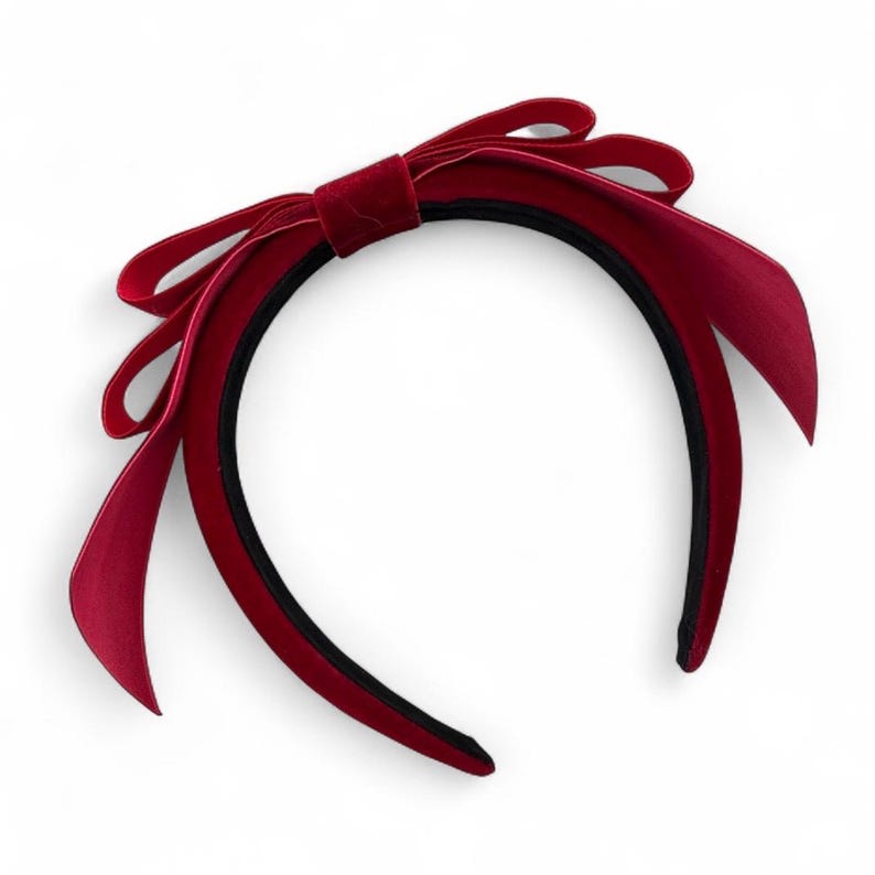 May include: A deep red velvet headband featuring a large, multi-layered bow detail on the side. The headband has a smooth, dark interior lining and a soft, textured exterior surface, creating a classic and structured appearance for this hair accessory.