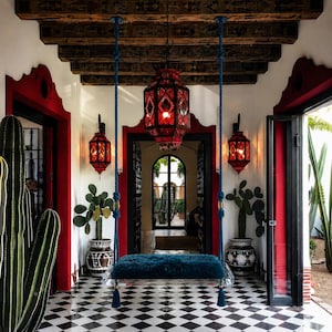 May include: A hanging swing seat with a dark teal faux fur cushion suspended by thick blue ropes. The scene features a black and white checkered floor, red door frames, three red geometric lanterns, and potted cacti in a bright entryway.